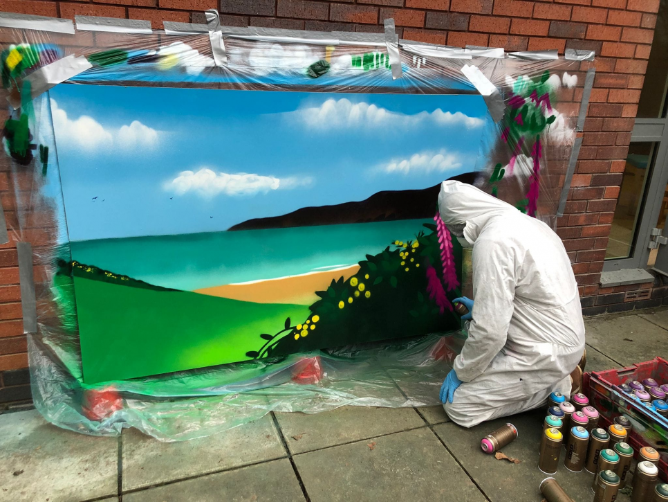 man kneeling spray painting a landscape onto a canvas