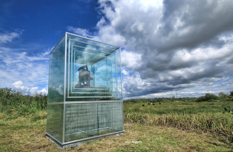 Art installation featuring a bell inside a transparent glass box, set in a grassy field under a partly cloudy sky.